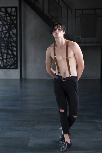Handsome shirtless dark haired male model in ripped jeans and suspenders in a dark room