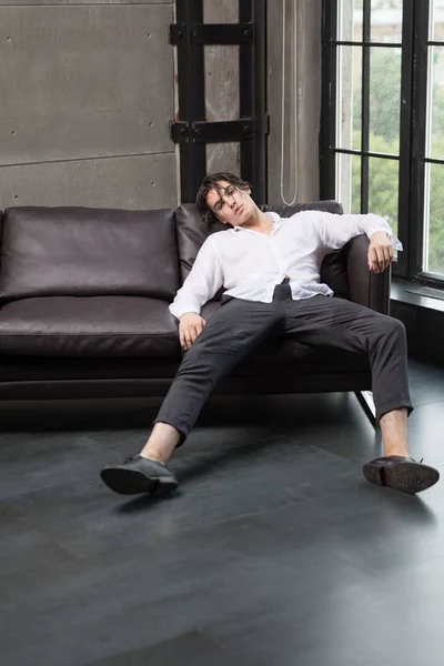 Şık yakışıklı delikanlı tüm beyaz gömlek ve rahat kameraya bakarak yaymak bacakları ile windows yakınındaki bir kanepede oturuyor gri pantolon ıslak siyah saçlı
