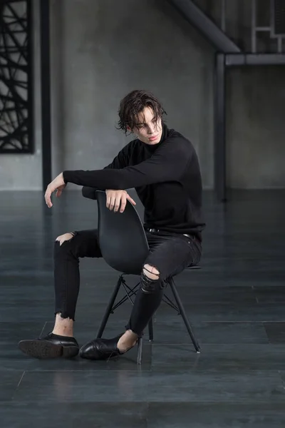 Şık yakışıklı delikanlı balıkçı yaka ve karanlık bir odada bir omuz üzerinden seyir çapraz kolları olan bir iskemlede yırtık kot pantolon ile tüm siyah kıyafeti ıslak siyah saçlı