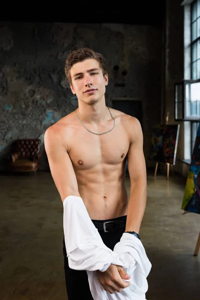 Handsome young man with brown hair teasing by undressing, taking off white shirt, revealing perfect body, six pack, muscle stomach at a trendy interior professional photostudio