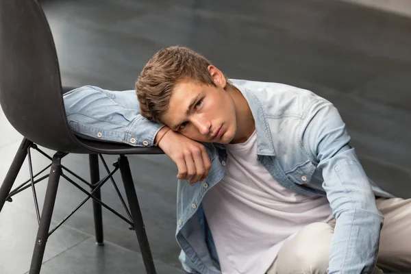 Handsome young male model sitting on a floor leaning on a chair looking at the camera