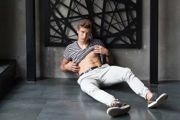 Handsome young male model in stripes t-shirt and white pants showing off six pack abs at modern interior photostudio