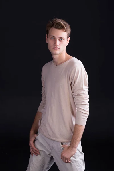 Handsome male model with blue eyes and light curlie hair in beige sweater and white pants on a dark background