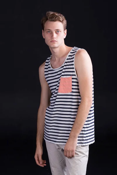 Handsome young man with blue eyes in a casual t-shirt with stripes and white trousers on a dark background