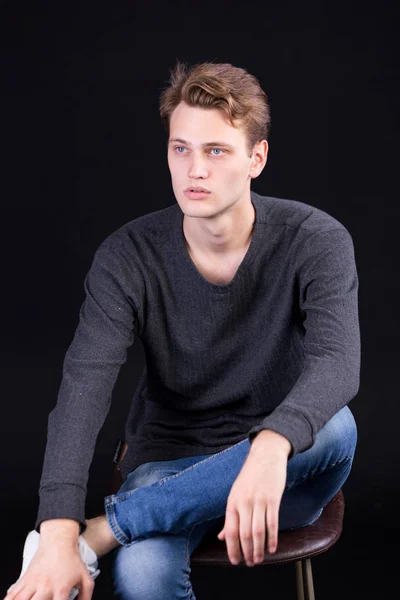 Handsome young man with blue eyes in a casual grey sweater with long sleeves and blue jeans sitting on a dark background