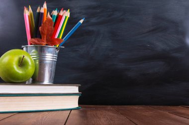 Ahşap masada kitap, yeşil elma, renkli kalemler ve sonbahar yaprağı yığını. Arka planda kopya alanı olan sınıf tahta. Eğitim, okul konseptine geri dön