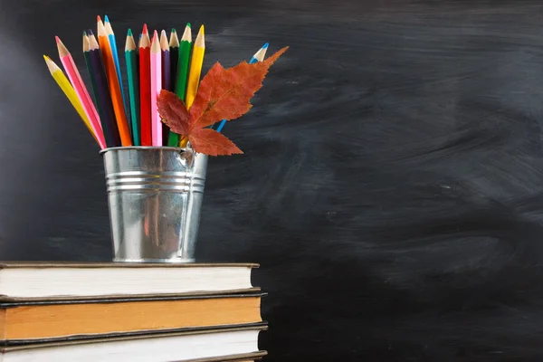 Colored pencils and fall leaf on stack of books. Classroom blackboard with copy space in background. Education, back to school concept