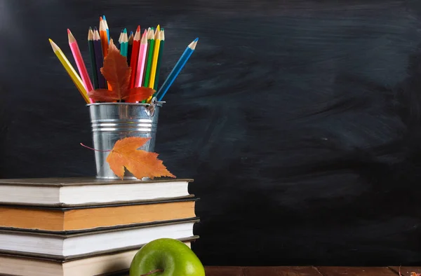 Colored pencils and fall leaves on stack of books. Green apple on wooden desk.  Classroom blackboard with copy space in background. Education, September, back to school concept