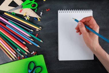 Boş bir defter sayfasına insan eli yazısı. Kalemler, yeşil defter, makas, raptiye, tahta zemin üzerinde cetvel. Üst görünüm, kopyalama alanı, düz yatış. Okul malzemeleri ya da ofis kırtasiyesi konsepti