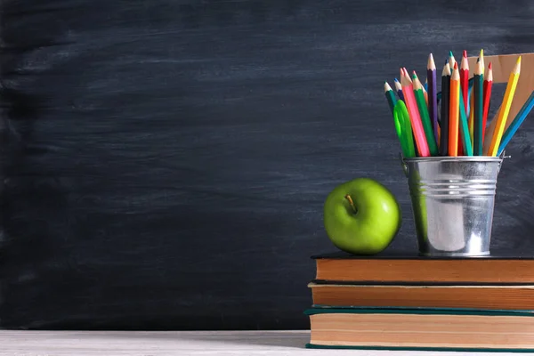 Bright green apple and pencils pot on stacked books, school supplies on white desk with blackboard texture in background. Side view, copy space, close-up. Learning, education concept