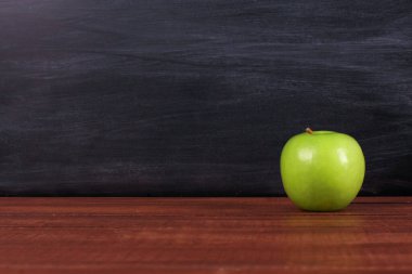 Kahverengi masanın üzerinde arka planda karatahta deseni olan tek bir yeşil elma. Uzayı kopyala, yakın plan. Okul atıştırmalıkları veya organik gıda konsepti