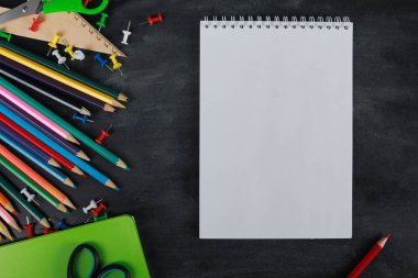 Defterin beyaz sayfası, metin için kopyalama alanı. Renkli kalemler, yeşil defter, makas, raptiyeler, tahta zemin üzerinde cetvel. Üst manzara, düz yatış. Okul malzemeleri ya da ofis kırtasiyesi konsepti