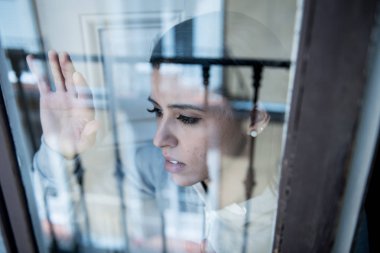Young beautiful depressed unhappy latin woman looking worried and sad through the window at home. feeling worthless and in pain. closeup. Depression and crisis concept clipart