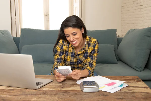 Happy attractive latin woman calculating home finances, accounting costs, charges, taxes, mortgage and paying bills at home using calculator and laptop looking cheerful and relax