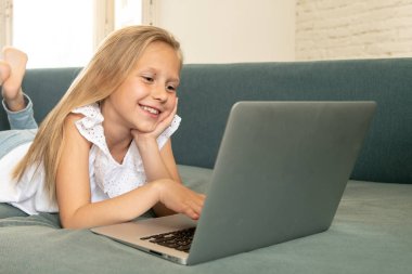Çok güzel oynayan ve gülümseyen ve evde kanepede uzanmış dizüstü bilgisayarda internet sörf küçük sarışın kız sevimli şirin. Dijital teknoloji, internet kullanımı ve yaşam tarzı kavramı