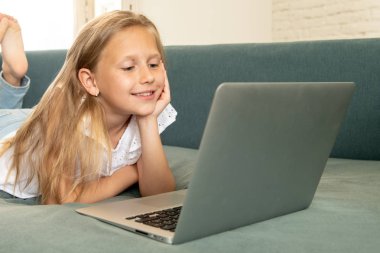 Çok güzel oynayan ve gülümseyen ve evde kanepede uzanmış dizüstü bilgisayarda internet sörf küçük sarışın kız sevimli şirin. Dijital teknoloji, internet kullanımı ve yaşam tarzı kavramı