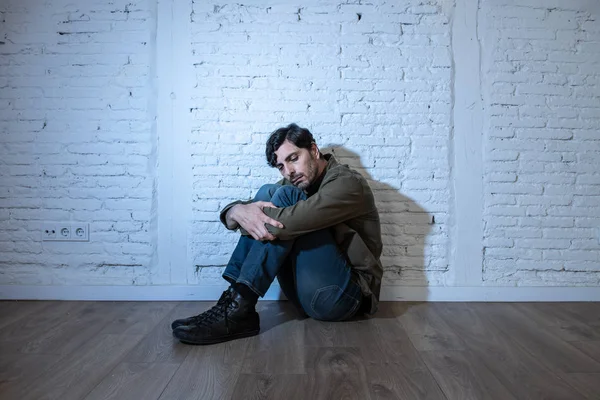 young depressed man sitting against a white wall at home with a shadow on the wall feeling miserable, lonely and sad in mental health depression concept