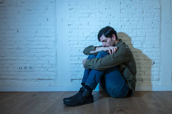 young depressed man sitting against a white wall at home with a shadow on the wall feeling miserable, lonely and sad in mental health depression concept