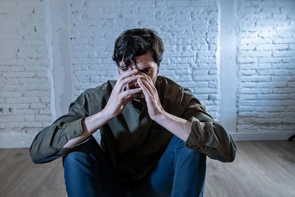 young depressed man sitting against a white wall at home with a shadow on the wall feeling miserable, lonely and sad in mental health depression concept