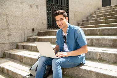 Genç yakışıklı modern adam açık merdivenlerde otururken dizüstü bilgisayar kullanan akıllı rahat kıyafet. Öğrenci ya da dalga köpüğü enterne açık havada serbest meslek