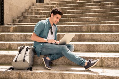 Genç yakışıklı modern adam açık merdivenlerde otururken dizüstü bilgisayar kullanan akıllı rahat kıyafet. Öğrenci ya da dalga köpüğü enterne açık havada serbest meslek