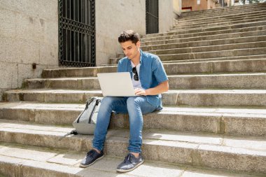 Genç yakışıklı modern adam açık merdivenlerde otururken dizüstü bilgisayar kullanan akıllı rahat kıyafet. Öğrenci ya da dalga köpüğü enterne açık havada serbest meslek