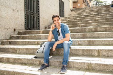 Merdivenler açık havada üzerinde oturan, cep telefonu üzerinde konuşurken, mutlu ve heyecanlı seyir çekici genç adam. Kentsel arka plan. İletişim teknolojisi kavramı.