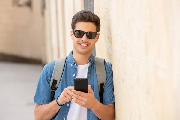 Outdoor shot of young male student in jeans denim clothes standing at street of modern European city and texting on smartphone