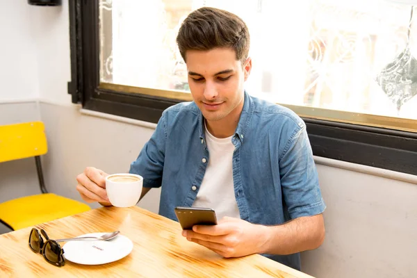 若い男性のフリーランサー座って自然食カフェでハンサムな男のコーヒー ショップで休憩中にテキスト メッセージを読む携帯電話のチャット — ストック写真