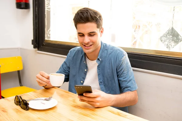 若い男性のフリーランサー座って自然食カフェでハンサムな男のコーヒー ショップで休憩中にテキスト メッセージを読む携帯電話のチャット — ストック写真
