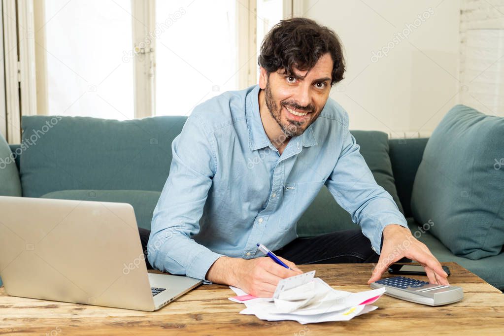 Happy attractive latin man calculating home finances, accounting costs, charges, taxes, mortgage and paying bills at home using calculator and laptop looking cheerful and relax at the camera