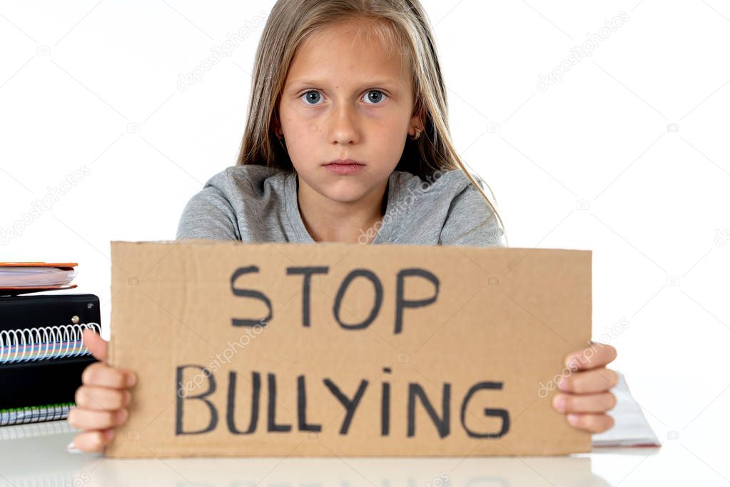 young cute caucasian blonde blue eyed schoolgirl scared and stressed holding paper with text stop bullying  asking for help sitting at school desk alone.  victim children bullied abuse concept
