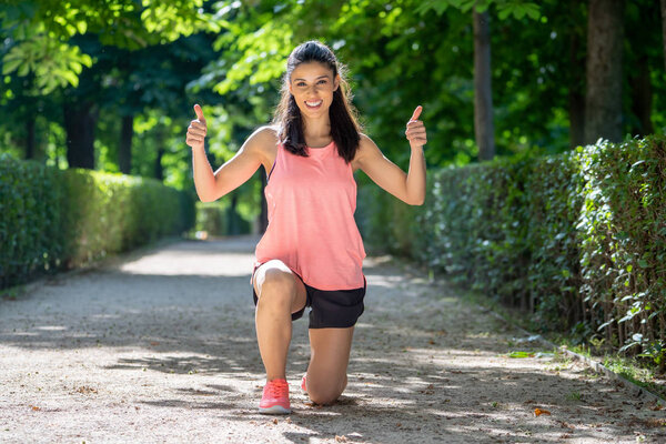 Summer portrait of sportive latin girl in pink tank top showing a thumb up cheerfully smiling and looking at camera working outside in a green park in fitness concept