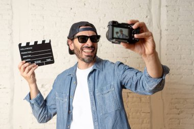 holding hollywood kayrak Yönetim Kurulu ile video fotoğraf makinesi tripod üzerinde tuğla duvar arka planda internet blog ve blogger kavramı karşı hippi tarzı rahat kıyafetler genç çekici adam.