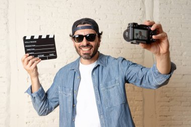 holding hollywood kayrak Yönetim Kurulu ile video fotoğraf makinesi tripod üzerinde tuğla duvar arka planda internet blog ve blogger kavramı karşı hippi tarzı rahat kıyafetler genç çekici adam.