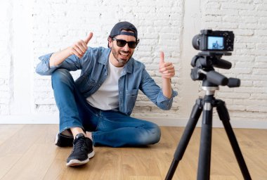 çekici bir genç adam rahat kıyafetler şapka sakallı hippi tarzı video kamera tripod karşı tuğla duvar arka planda internet blog ve blogger kavramı üzerinde. Başparmak göstermek yukarıya adam