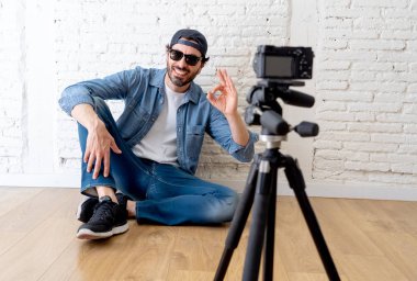 çekici bir genç adam rahat kıyafetler şapka sakallı hippi tarzı video kamera tripod karşı tuğla duvar arka planda internet blog ve blogger kavramı üzerinde. Tamam gösterilen adam 