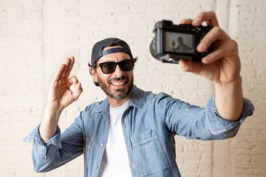 genç çekici adam hippi tarzı modern bir görünüm kendi kendine portre çekim ya da yazmak video internet blog ve blogger kavramında fotoğraf kamera tutarak rahat kıyafetler giyiyor. Tamam gösterilen adam