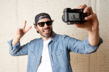 genç çekici adam hippi tarzı modern bir görünüm kendi kendine portre çekim ya da yazmak video internet blog ve blogger kavramında fotoğraf kamera tutarak rahat kıyafetler giyiyor. Adam gösteren zafer işareti 