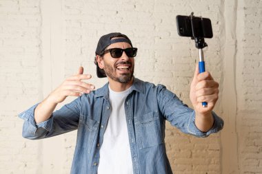 young attractive man in casual clothes, hat in  hipster style holding selfie stick mobile phone shooting self portrait photo or recording video.  internet blog and blogger concept. clipart