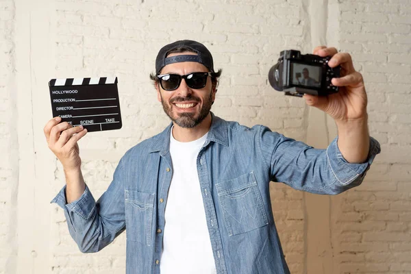 holding hollywood kayrak Yönetim Kurulu ile video fotoğraf makinesi tripod üzerinde tuğla duvar arka planda internet blog ve blogger kavramı karşı hippi tarzı rahat kıyafetler genç çekici adam.