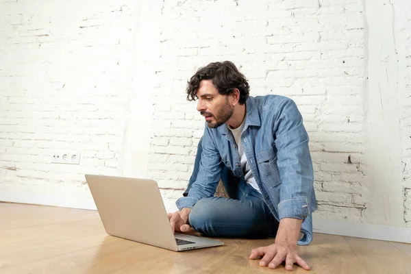 T恤衫和牛仔裤的拉丁男士肖像坐在白色砖墙上 手持笔记本电脑 微笑着看着媒体 从家庭概念的时髦自由职业者工作 — 图库照片