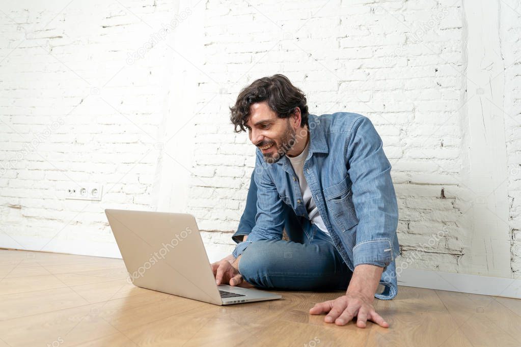 portrait of latin man dressed in T-shirt blue shirt and jeans sitting against white brick wall holding laptop and watching media with happy smile. hipster freelance working from home concept.