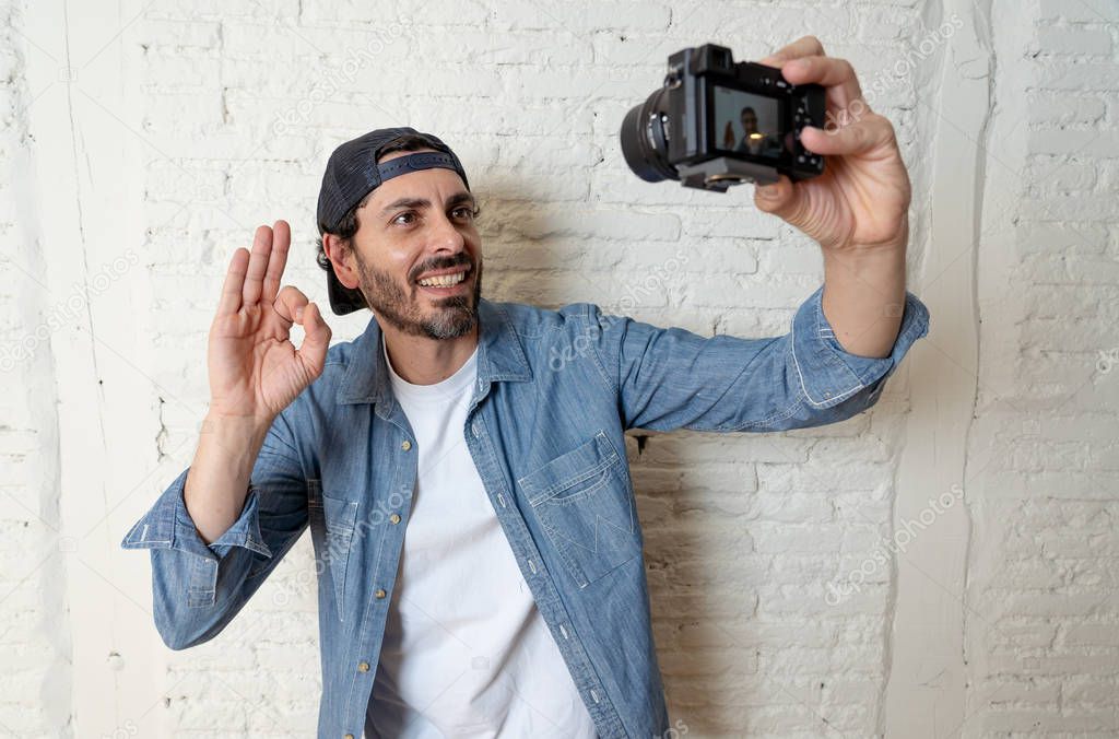 young attractive man wearing casual clothes  in hipster style modern look holding photo camera shooting self portrait  or recording video in internet blog and blogger concept. Man showing ok