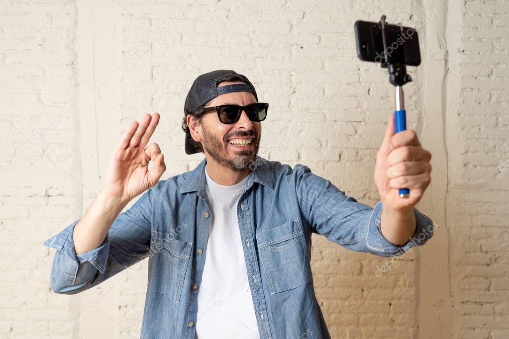 young attractive man in casual clothes, hat in  hipster style holding selfie stick mobile phone shooting self portrait photo or recording video.  internet blog and blogger concept. Man showing  ok