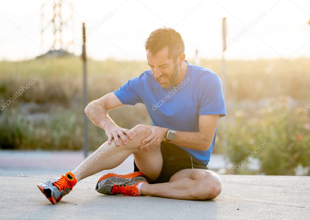young sport man with strong athletic legs holding knee with his hands in pain after suffering muscle injury during a running workout training in asphalt road in muscular or ligament injury concept.