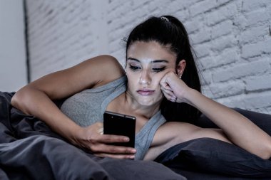 Genç güzel Latin kadın yatakta geç konuşuyor ve cep telefonuyla geceleri sıkılmış ve Internet iletişim aşırı kavramı ve akıllı telefon bağımlılığı akıllı cep telefonu kullanarak yorgun.