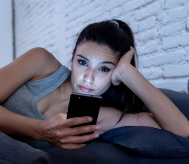 Genç güzel Latin kadın yatakta geç konuşuyor ve cep telefonuyla geceleri sıkılmış ve Internet iletişim aşırı kavramı ve akıllı telefon bağımlılığı akıllı cep telefonu kullanarak yorgun.