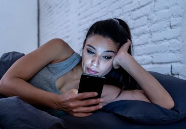 Genç güzel Latin kadın yatakta geç konuşuyor ve cep telefonuyla geceleri sıkılmış ve Internet iletişim aşırı kavramı ve akıllı telefon bağımlılığı akıllı cep telefonu kullanarak yorgun.