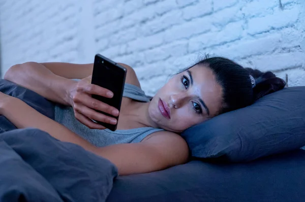 話していると 退屈やインターネット通信酷使概念とスマート フォン中毒でスマートな携帯電話を使用しての疲れの携帯電話を使用して深夜にベッドの上若いラテン系美人 — ストック写真
